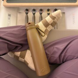 Sleek brown stainless steel water bottle balancing on the legs of someone sitting on a reformer pilates machine